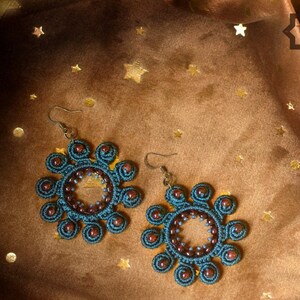 May include: A pair of teal blue and brown crocheted earrings with a circular design. The earrings are on a brown background with gold stars.