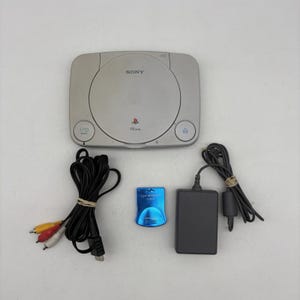 May include: A gray PlayStation PSone console with the Sony logo, power and eject buttons, and a circular disc tray. Includes a power adapter, AV cables, and a blue memory card.