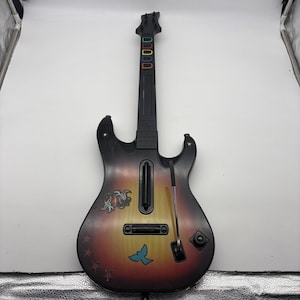 May include: A black and sunburst-colored plastic video game guitar controller featuring five colored fret buttons on the neck, a strum bar, a whammy bar, and decorative decals including a blue bird, stars, and a stylized graphic on the body.