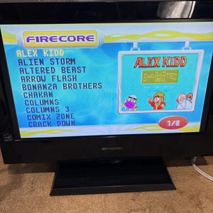 May include: A black Emerson TV displaying a retro video game menu. The screen shows a list of games including "ALEX KIDD" and "ALIEN STORM." The game "ALEX KIDD" is highlighted with colorful graphics. The screen displays "1/8" at the bottom.