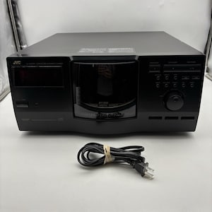 May include: Black JVC CD player with a digital display and various control buttons. The unit has a disc tray and a power cord. The front panel features the JVC logo. A black power cord is coiled in front of the player.