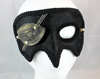 May include: A black masquerade mask with a gold eye patch and black and white trim. The mask is on a white mannequin head.