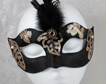 May include: A black masquerade mask with gold and silver sequins and a black feather. The mask has a jeweled embellishment in the center.