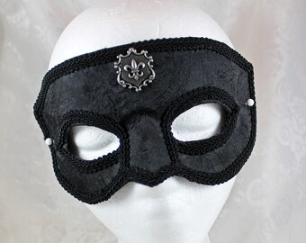 May include: A black masquerade mask with a silver fleur-de-lis embellishment. The mask has a black velvet texture and a black satin trim.