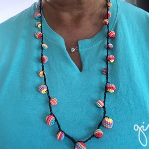 May include: A handmade necklace featuring colorful, crocheted ball beads in shades of pink, yellow, blue, and orange. The beads are strung on a black cord. The necklace is worn against a blue shirt.