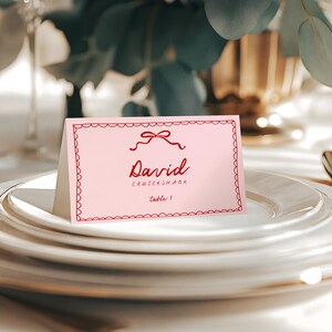 May include: A pink place card with a red scalloped border and bow design. The card displays the name "David" in cursive, with the last name "Cruickshank" below, and "table 1" at the bottom. The card is set on a stack of white plates.