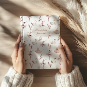 Può includere: Un diario a tema balletto. La copertina presenta un motivo di ballerine, cigni e scarpette da ballo nei toni del rosa e del bianco. Il testo "Charlotte's Journal" è in una cornice decorativa.