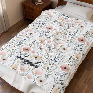 Puede incluir: Una manta blanca con un estampado floral de flores rosas y beige y hojas verdes. La palabra "Sarah" está escrita en cursiva negra en la manta. La manta está sobre una cama con una almohada blanca.