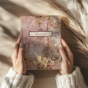 May include: A journal with a pink and gold floral design, held by a person wearing a cream-colored sweater. The journal cover features the name "Charlotte" in a rectangular label. The design includes script and ornate gold embellishments.