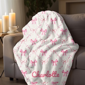 May include: A white plush blanket with a pattern of pink bows and ribbons. The name "Charlotte" is printed in pink script at the bottom. The blanket is draped over a gray sofa.