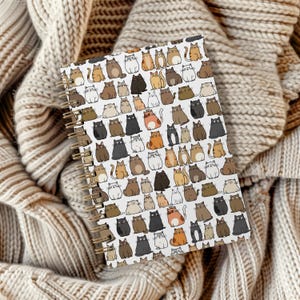 May include: A white spiral bound notebook with a colorful pattern of cartoon cats. The cats are in various poses and colors, including orange, brown, black, and white.
