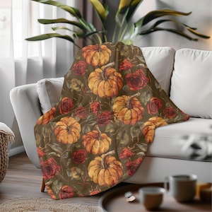 Op de afbeelding: Een decoratieve deken met een herhalend patroon van oranje pompoenen en rode rozen op een donkerbruine achtergrond. De deken is over een witte bank gedrapeerd, wat een warme, herfstachtige sfeer creëert.