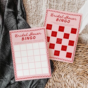 Puede incluir: Dos tarjetas de bingo de despedida de soltera de color rosa con texto y bordes rojos. La tarjeta superior tiene una lista de regalos para marcar, incluyendo toallas de cocina, cuchillos para pasteles, tazas de café, tazones para mezclar, y más. La tarjeta inferior está en blanco para que los jugadores marquen los regalos a medida que se abren.