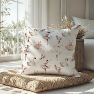 May include: A white cushion with a ballet design. It features illustrations of ballerinas, tutus, pointe shoes, and swans in pink and white. The name "Victoria" is written on the cushion in a cursive font.