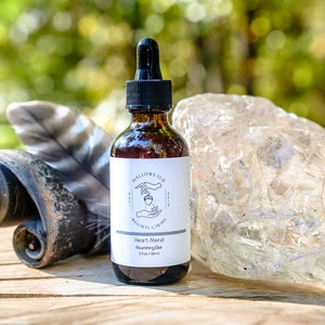 May include: A brown glass bottle with a dropper top and a white label that reads "Hallowild Wellness & Grace" and "Heart Mind Nourishing Elixir 2 fl oz / 60 ml". The bottle is sitting on a wooden surface with a feather and a clear crystal rock.