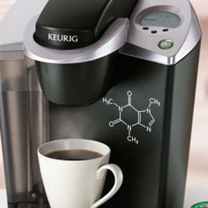 Op de afbeelding: Een zwarte en zilveren Keurig koffiezetapparaat voor &eacute;&eacute;n kopje met een witte mok gevuld met koffie. Het koffiezetapparaat heeft een digitaal display en een cafe&iuml;nemolecuulafbeelding. Verschillende K-Cup cups zijn zichtbaar.