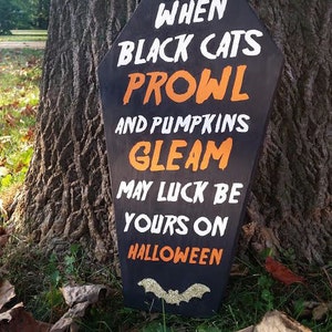 May include: Black coffin-shaped Halloween sign with white and orange text: "When black cats prowl and pumpkins gleam, may luck be yours on Halloween." A glittery bat is at the bottom.