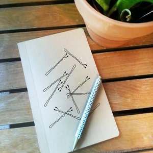 May include: A beige notebook with a pen and bobby pins on a wooden surface. The pen has a white and silver dotted pattern. The bobby pins are drawn on the notebook. A potted plant is in the background.