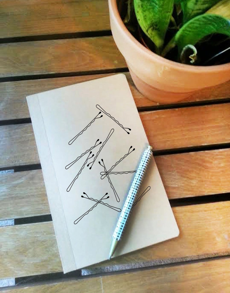 May include: A beige notebook with a pen and bobby pins on a wooden surface. The pen has a white and silver dotted pattern. The bobby pins are drawn on the notebook. A potted plant is in the background.