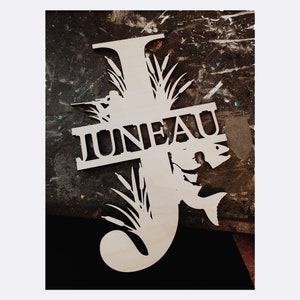 May include: Wooden cutout in the shape of the letter "J" with the word "JUNEAU" cut out. The design includes silhouettes of a fisherman, a fish, and reeds. The cutout is made of light-colored wood.