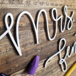 May include: Wooden sign with the words "Mrs. [Name]" in white cursive lettering. The background is a dark wood with ruler markings. A purple eraser and a yellow pencil are visible.