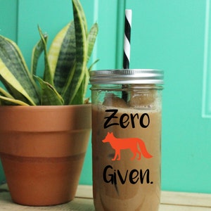Puede incluir: Un tarro de cristal transparente lleno de una bebida marrón y cubitos de hielo, coronado con una tapa plateada y una pajita a rayas blancas y negras. El tarro tiene la inscripción "Zero Given" con un zorro naranja. Una planta en maceta está en el fondo.