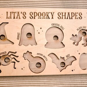 May include: Wooden Halloween puzzle featuring cut-out shapes: a witch's hat, ghost, crystal ball, black cat, spider, potion bottle, bat, and pumpkin. The text "LITA'S SPOOKY SHAPES" is engraved on the top of the puzzle.