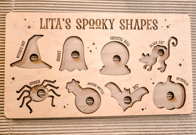 May include: Wooden Halloween puzzle featuring cut-out shapes: a witch's hat, ghost, crystal ball, black cat, spider, potion bottle, bat, and pumpkin. The text "LITA'S SPOOKY SHAPES" is engraved on the top of the puzzle.