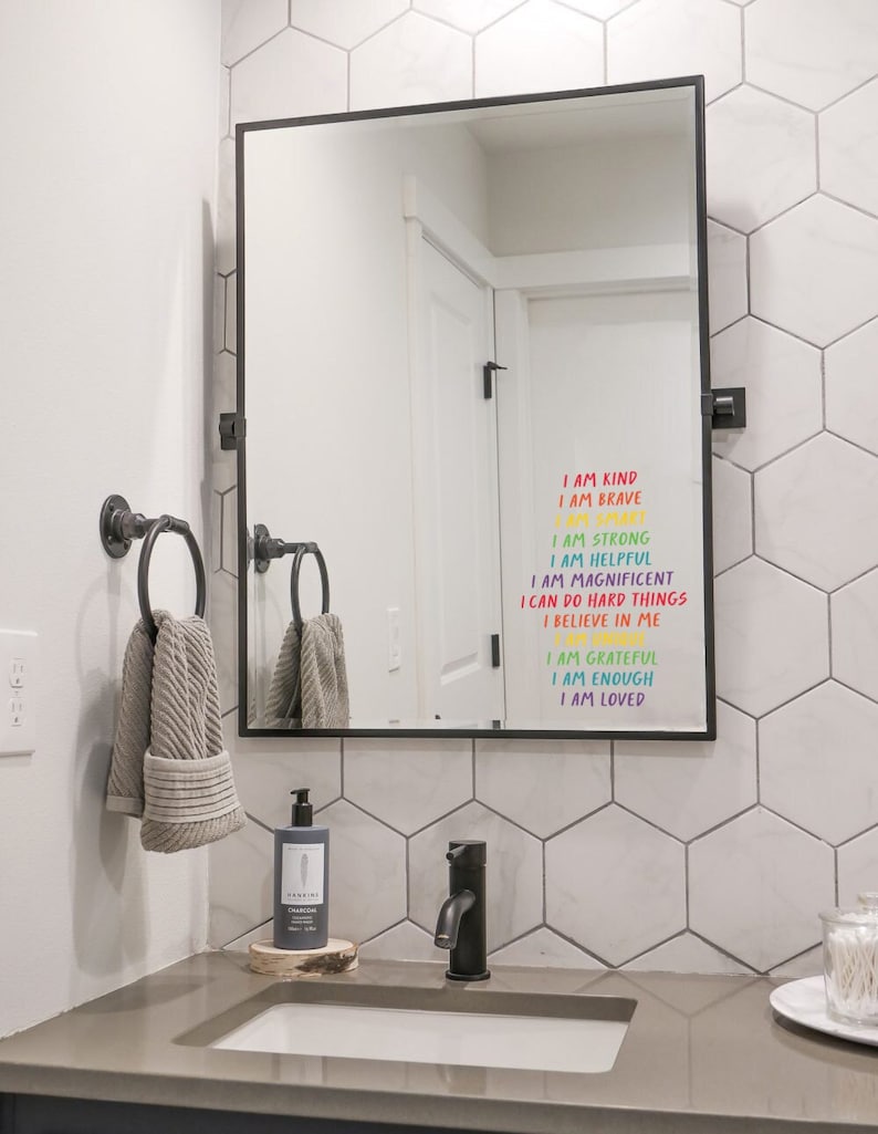 May include: A black-framed mirror hangs above a bathroom sink, reflecting a white door. The mirror has colorful text that reads "I AM KIND, I AM BRAVE, I AM STRONG, I AM HELPFUL, I AM MAGNIFICENT, I CAN DO HARD THINGS, I BELIEVE IN ME, I AM GRATEFUL, I AM ENOUGH, I AM LOVED."