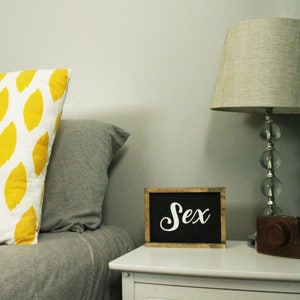 May include: A bedroom scene with a white nightstand, a lamp, and pillows. A small chalkboard sign with the word "Sex" in white script sits on the nightstand. A decorative pillow with a yellow leaf pattern rests on the bed.