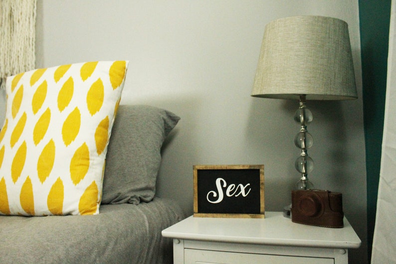 May include: A bedroom scene with a white nightstand, a lamp, and pillows. A small chalkboard sign with the word "Sex" in white script sits on the nightstand. A decorative pillow with a yellow leaf pattern rests on the bed.