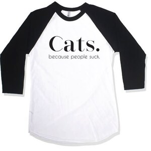 以下が含まれることがあります： 「Cats. because people suck」のテキストが入った白と黒のベースボールTシャツ。シャツは白いボディと黒い袖が特徴です。テキストは太字のサンセリフ体です。