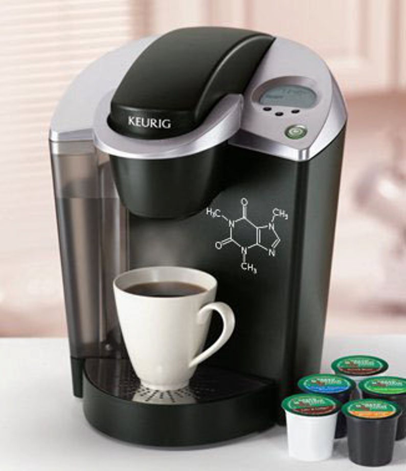 Op de afbeelding: Een zwarte en zilveren Keurig koffiezetapparaat voor &eacute;&eacute;n kopje met een witte mok gevuld met koffie. Het koffiezetapparaat heeft een digitaal display en een cafe&iuml;nemolecuulafbeelding. Verschillende K-Cup cups zijn zichtbaar.