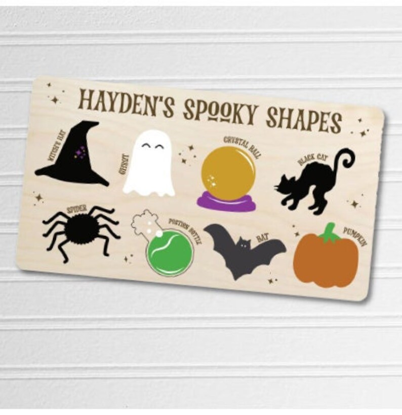 May include: Wooden Halloween puzzle board with the text "HAYDEN'S SPOOKY SHAPES". It features illustrations of a witch's hat, ghost, crystal ball, black cat, spider, potion bottle, bat, and pumpkin.