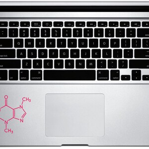 Op de afbeelding: Een zilveren laptop met een zwart toetsenbord en een roze sticker van een cafe&iuml;nemolecuul op de polssteun. De laptop is open en toont het toetsenbord en het touchpad. De sticker is een gestileerde chemische structuur.