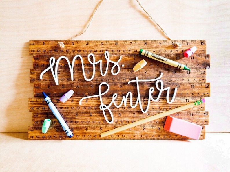 May include: Wooden sign with the name "Mrs. Fenter" in white script, decorated with school supplies like crayons, a pencil, and an eraser. The sign is made of wooden rulers and has a twine hanger.