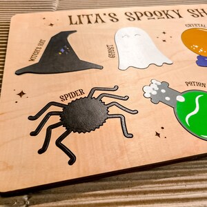 May include: Wooden board featuring "Lita's Spooky Shapes" with Halloween illustrations. Includes a witch's hat, ghost, crystal ball, spider, and potion bottle. The shapes are in black, white, orange, and green, on a light wood background.