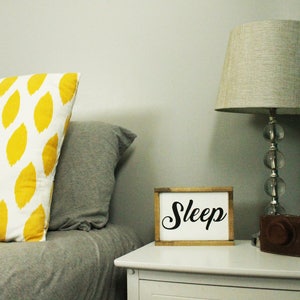 May include: A bedroom scene featuring a white nightstand with a lamp and a sign that says "Sleep". A decorative pillow with a yellow leaf pattern rests on a bed with a gray comforter. A brown leather camera case sits on the nightstand.