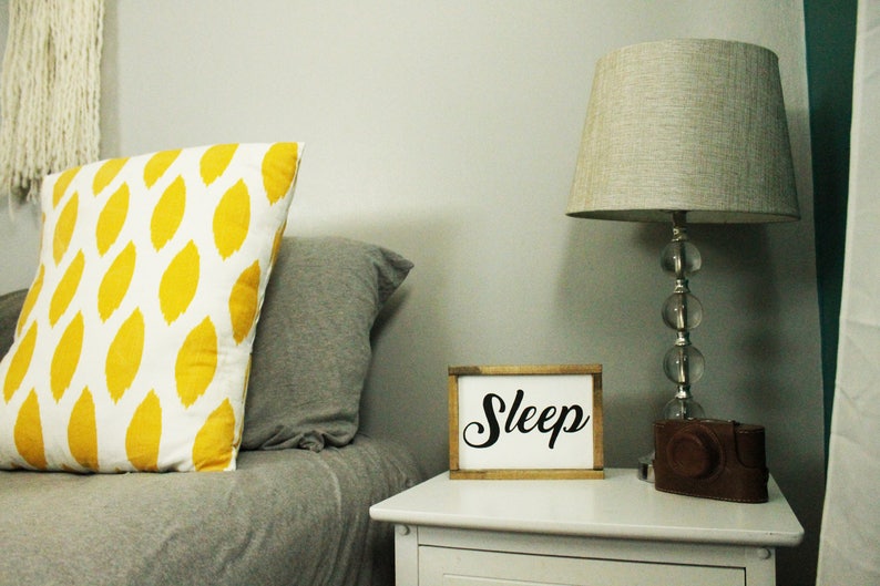 May include: A bedroom scene featuring a white nightstand with a lamp and a sign that says "Sleep". A decorative pillow with a yellow leaf pattern rests on a bed with a gray comforter. A brown leather camera case sits on the nightstand.