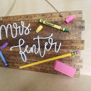 May include: A wooden sign with the name "Mrs. Fenter" in white script. The sign is designed to look like a ruler and includes a yellow pencil, a pink eraser, and colorful crayons. A perfect gift for a teacher.