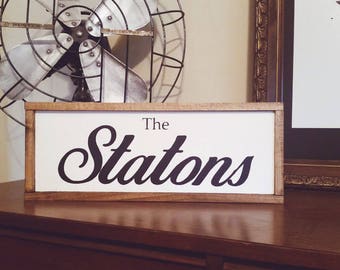 May include: A wooden framed sign with the words "The Statons" in a black, elegant script. The sign is set on a dark wooden surface, with a vintage fan and framed artwork in the background. The sign is a decorative home accent.