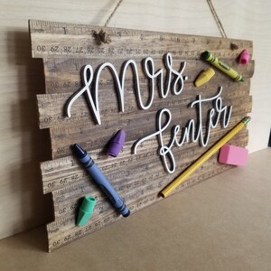 May include: Wooden sign with the name "Mrs. Fenter" in white script, decorated with colorful crayons, a pencil, and an eraser. The sign has a ruler design and is suspended by a twine string.