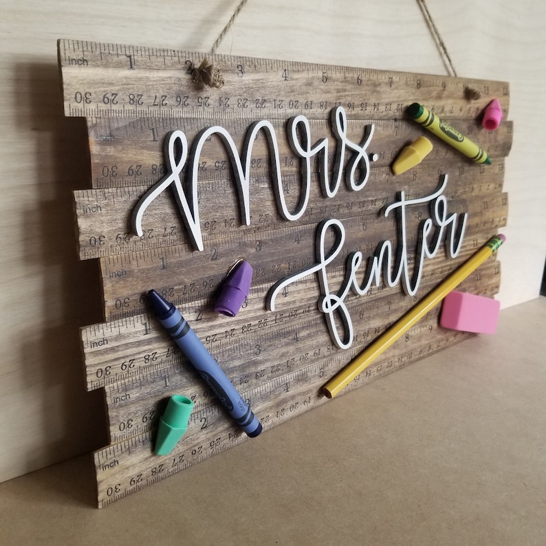 May include: Wooden sign with the name "Mrs. Fenter" in white script, decorated with colorful crayons, a pencil, and an eraser. The sign has a ruler design and is suspended by a twine string.