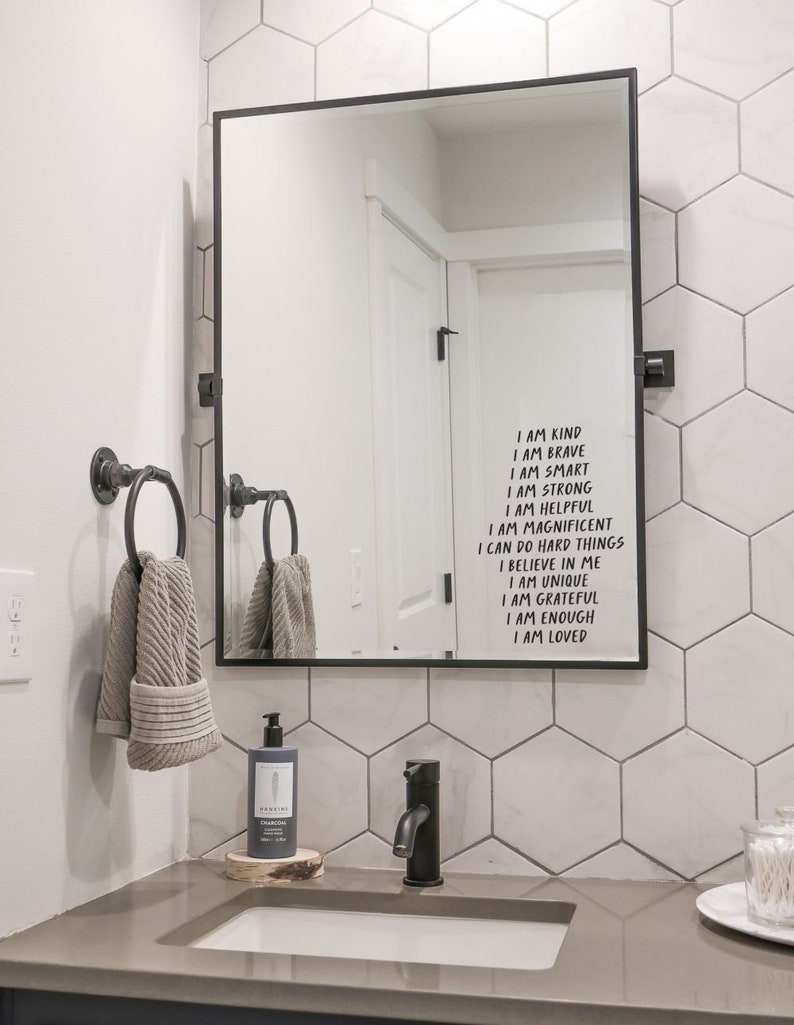 May include: A modern bathroom mirror with a black frame, displaying inspirational text. The mirror hangs above a gray countertop with a black faucet, soap dispenser, and a wooden coaster. Two gray towels hang on a towel rack.