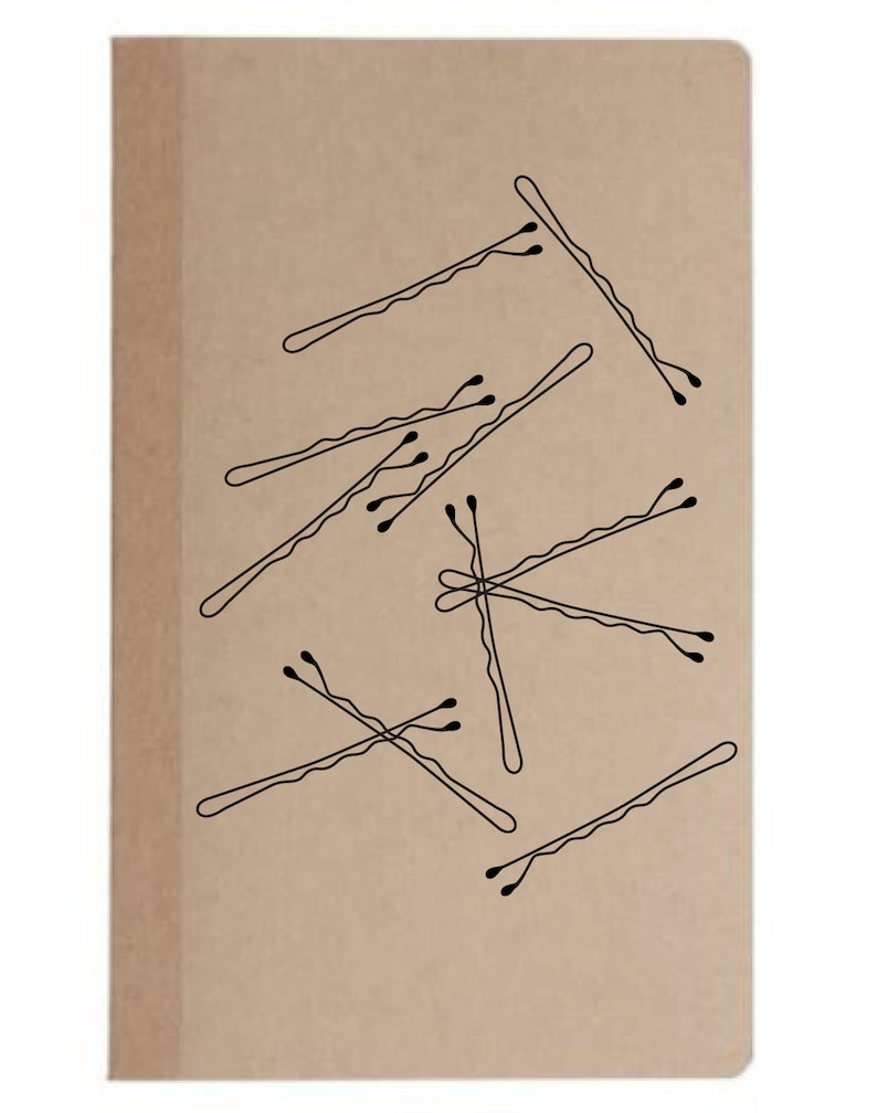 May include: A tan notebook with a collection of black bobby pins scattered across the cover. The bobby pins are drawn in a simple, minimalist style, with a wavy shape and small, rounded tips. The notebook has a plain, unadorned design.