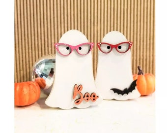 Può includere: Due decorazioni bianche a forma di fantasma con occhiali rosa e rossi e la parola "Boo" in arancione. Sono presenti anche un pipistrello nero e zucche arancioni, che suggeriscono un tema di Halloween.