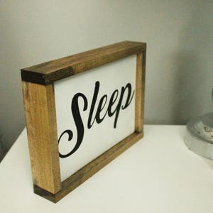 May include: Wooden framed sign with the word "Sleep" in black script on a white background. The sign is on a white surface next to a lamp and a brown leather case.