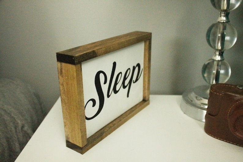 May include: Wooden framed sign with the word "Sleep" in black script on a white background. The sign is on a white surface next to a lamp and a brown leather case.