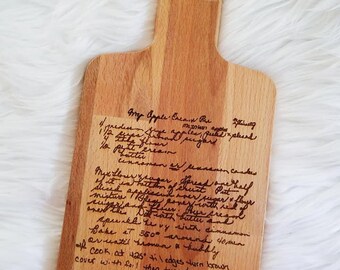 May include: A wooden cutting board with a handle and a hole for hanging. The board features handwritten text of a recipe for "Crisp Apple Cream Pie". The wood has a light brown color.