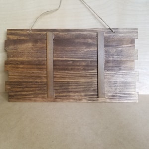 May include: A rectangular wooden sign with a dark brown stain. The sign is constructed from horizontal wooden planks of varying lengths, with two vertical support pieces. A twine loop is attached for hanging.